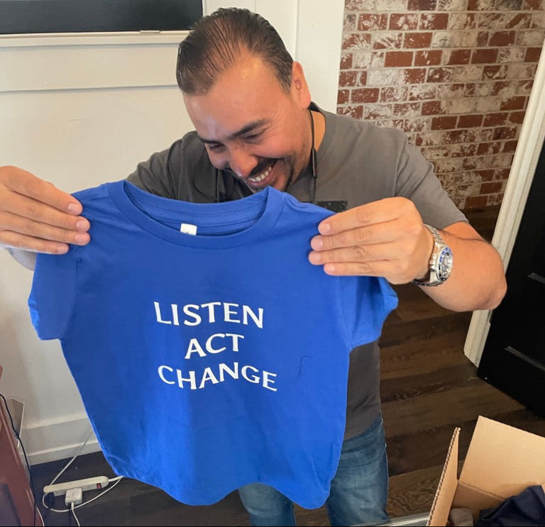 Man holding a blue t-shirt with 'Listen Act Change' text in a casual indoor setting.